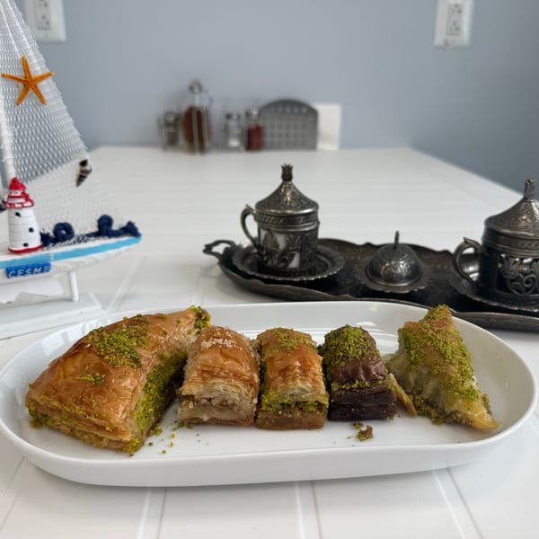 A white plate of pistachio-topped baklava next to decorative silver coffee cups and a sailboat.