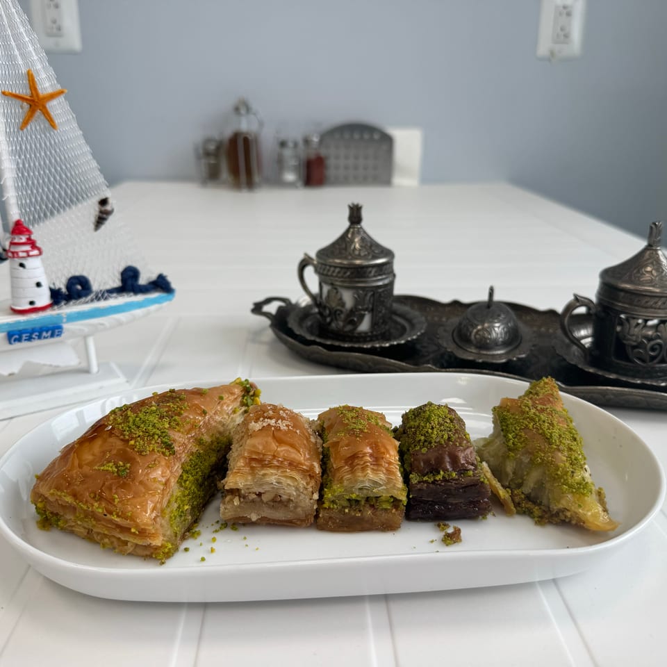 A white plate of pistachio-topped baklava next to decorative silver coffee cups and a sailboat.