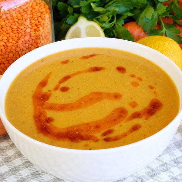 Bowl of lentil soup with red oil swirl, surrounded by lentils, onion, lemons, carrots, and parsley.